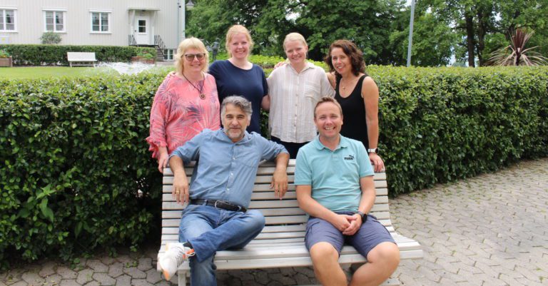 Seks personer avbildet foran en grønn hekk og en lys bygning