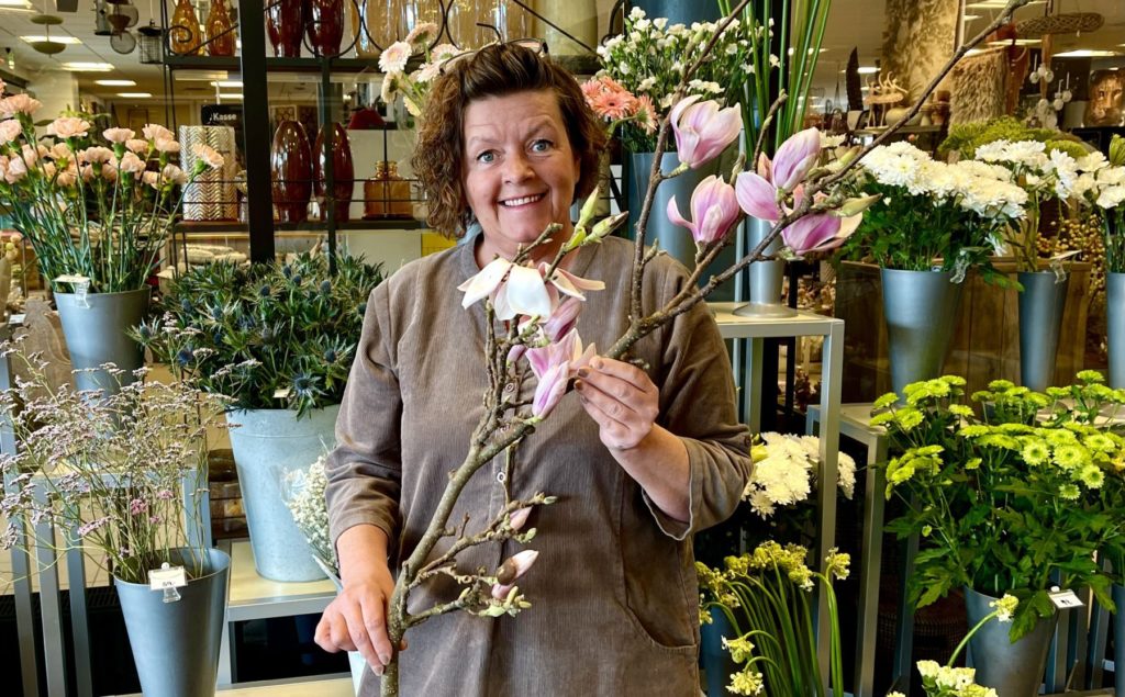 Dame med mørkt krøllete hår holder en grein med magnolia, inne i en blomsterforretning