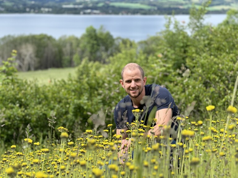 Mann som sitter på huk i en blomstereng, Mjøsa i bakgrunnen
