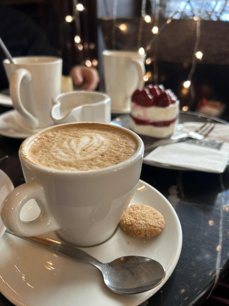 Nærbilde av hvit kaffekopp med cappuccino og en kake med bær i bakgrunnen