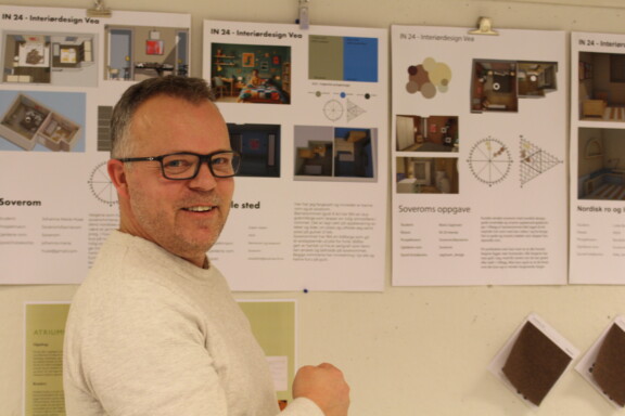 Mann med kort hår og briller står foran en oppslagstavle i et klasserom