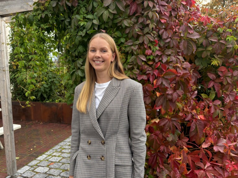 Dame med langt lys hår og dressjakke, avbildet ute foran en høstfarget villvin