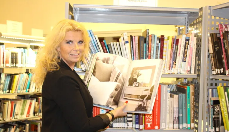 Dame stående i bibliotek med en interiørbok i hånden