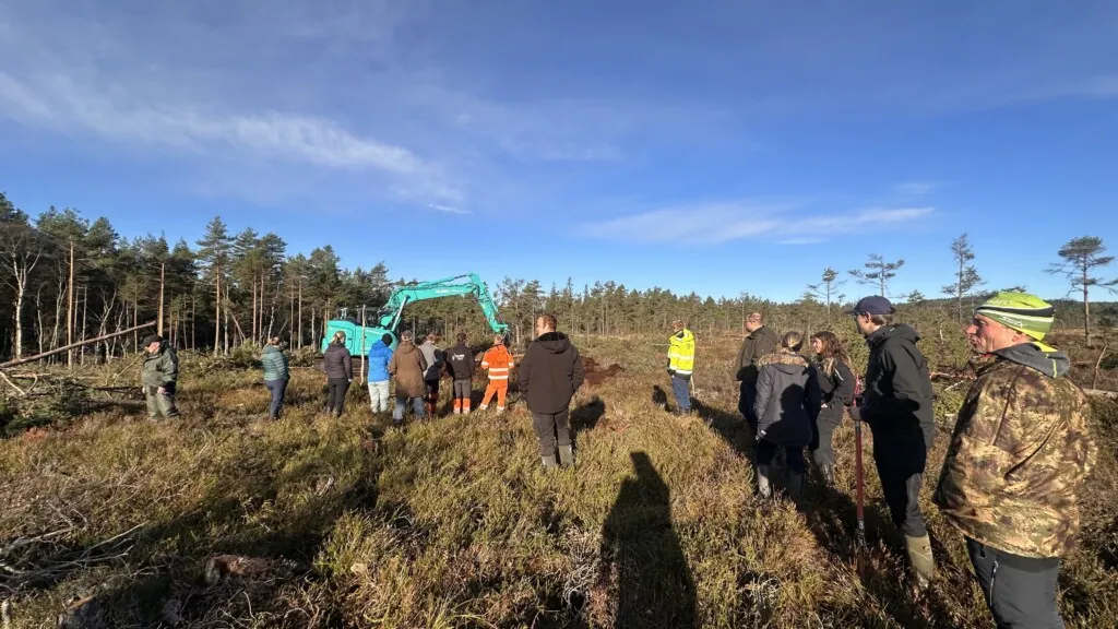 Turkis gravemaskin graver i myr og mange personer følger med