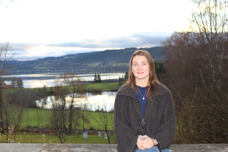 Dame med mørkt hår i grå genser sitter på en murkant med Mjøsa i bakgrunnen