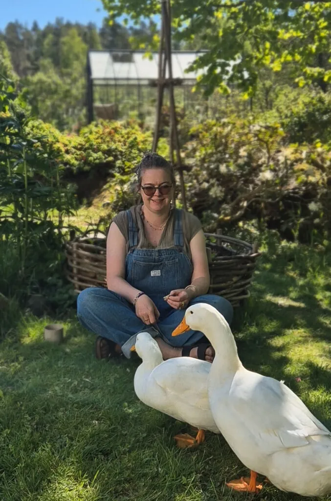 Kvinne iført snekkerbukse sitter i en hage og mater to gjess