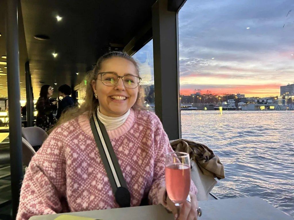 Smilende jente med briller, rosa strikket genser ombord i en båt på Donau, med Budapest i bakgrunnen