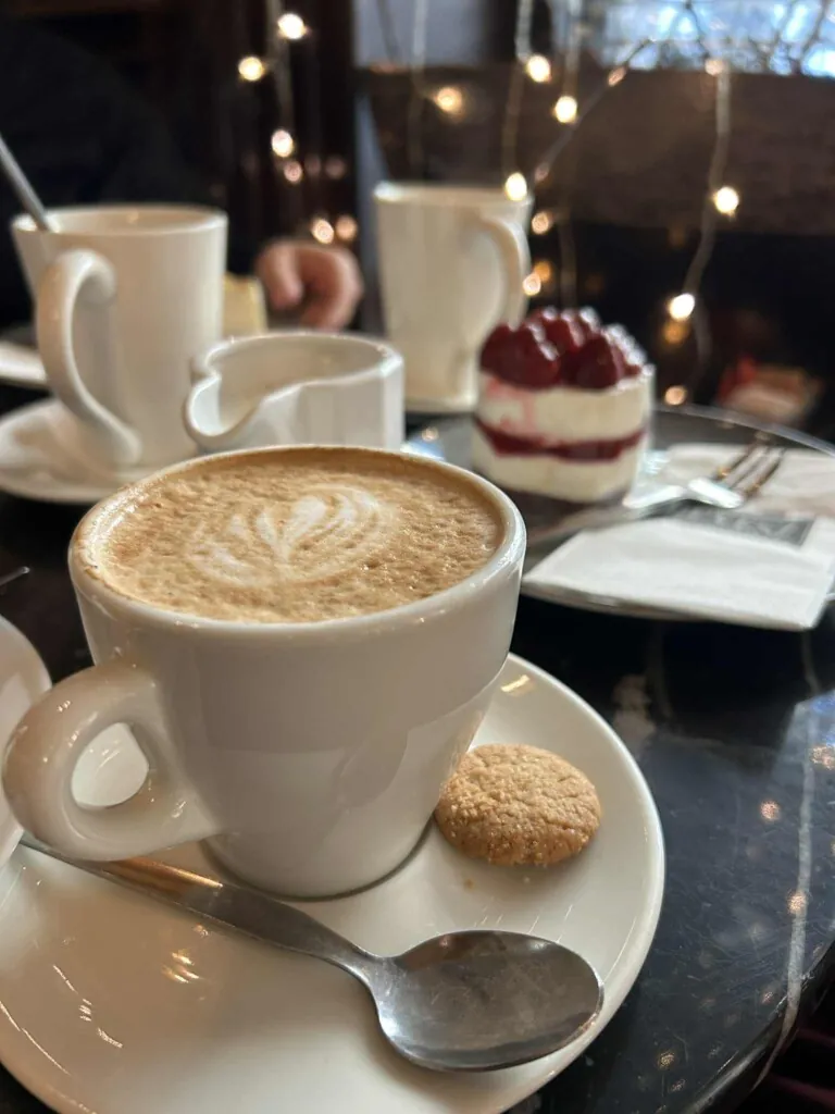 Nærbilde av hvit kaffekopp med cappuccino og en kake med bær i bakgrunnen