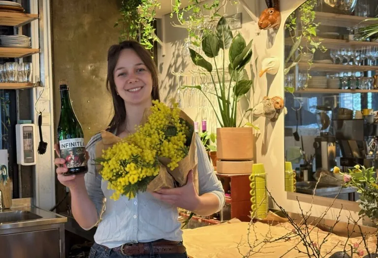 Jente med mørkt langt hår står inne i en blomsterbutikk og holder en flaske og en blomsterbukett