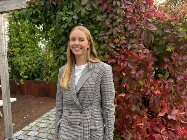Dame med langt lys hår og dressjakke, avbildet ute foran en høstfarget villvin
